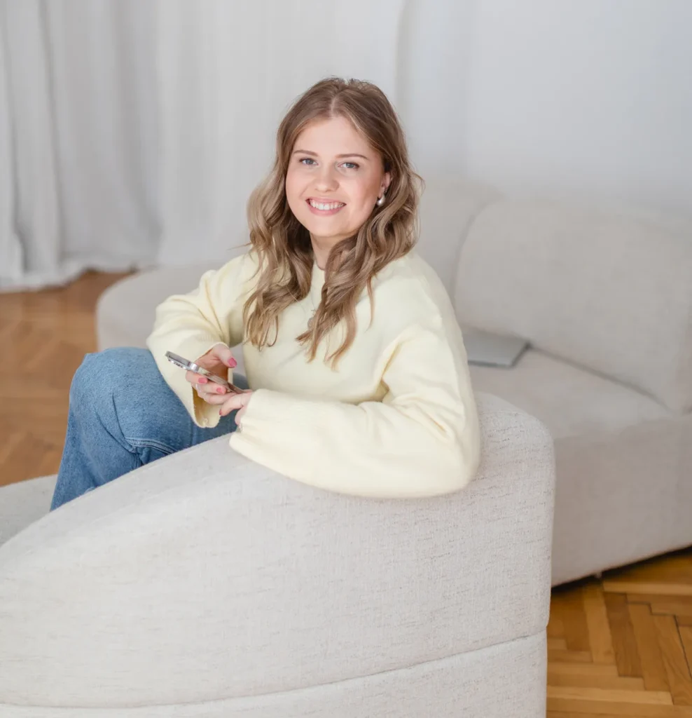 Katrin sitzt lächelnd in einem hellen Sessel und hält ein Smartphone in der Hand