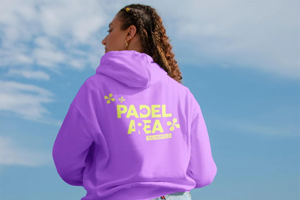 Person trägt einen gebrandeten Padel Area Tullnerfeld Hoodie in auffälligem Lila mit neongrünem Logo auf der Rückseite. Das Design verbindet Sport, Lifestyle und Markenidentität in einem modernen Merchandise-Produkt.